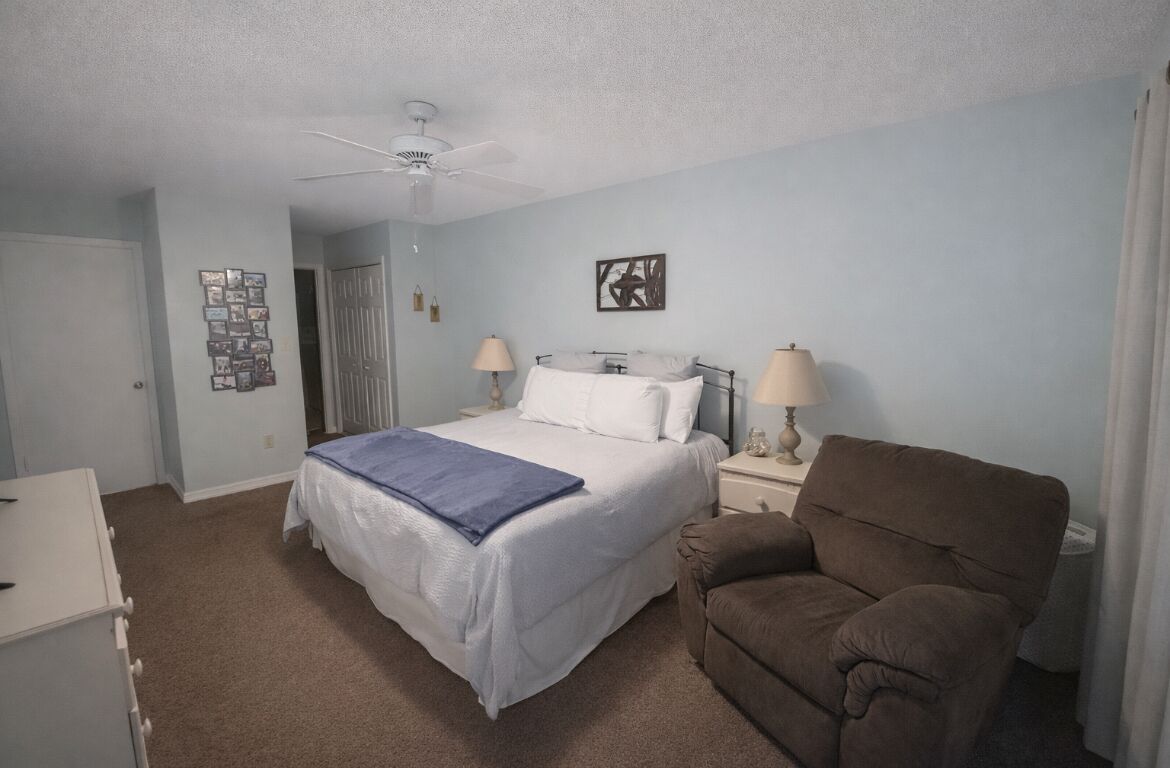 Une chambre à coucher avec un lit, deux lampes de chevet, un ventilateur de plafond, un fauteuil marron et un mur orné de tableaux encadrés.
