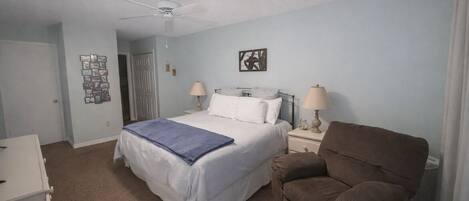 Une chambre à coucher avec un lit, deux lampes de chevet, un ventilateur de plafond, un fauteuil marron et un mur orné de tableaux encadrés.