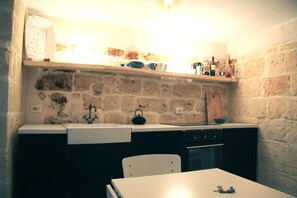 A kitchen with a stone wall, a white countertop, a sink, a kettle, and a wooden cutting board.