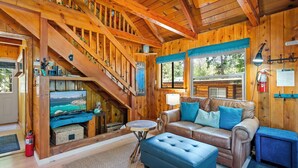 Ein Holzblockhaus-Innenraum mit Treppe, Ledersofa, Hocker und einem Fernseher, der eine Berglandschaft zeigt.