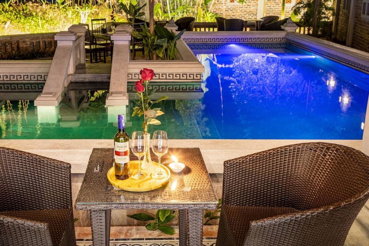 A dining table with wicker chairs, a bottle of wine, and a glass on a tray, overlooking a pool with a blue water feature.