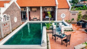 A swimming pool with a brick wall and a tiled roof, surrounded by outdoor seating and plants. - Paddy Home (Cam Pha)