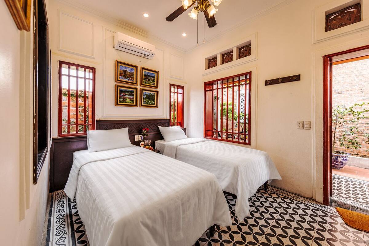A hotel room with two beds, a ceiling fan, an air conditioning unit, and a view of the outside through barred windows.