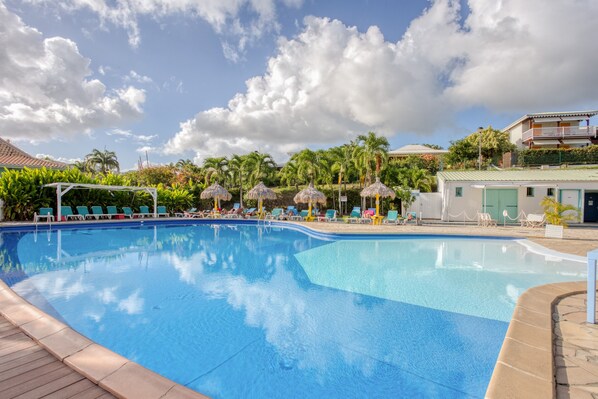 A swimming pool with lounge chairs and umbrellas, surrounded by palm trees and buildings.