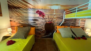 Een kamer met twee bedden, elk met een groene plaid en meerdere kussens. Op de muur bevindt zich een muurschildering van een strand met boten en een zonsondergang. Een houten kruk met een roze bloem staat tussen de bedden.