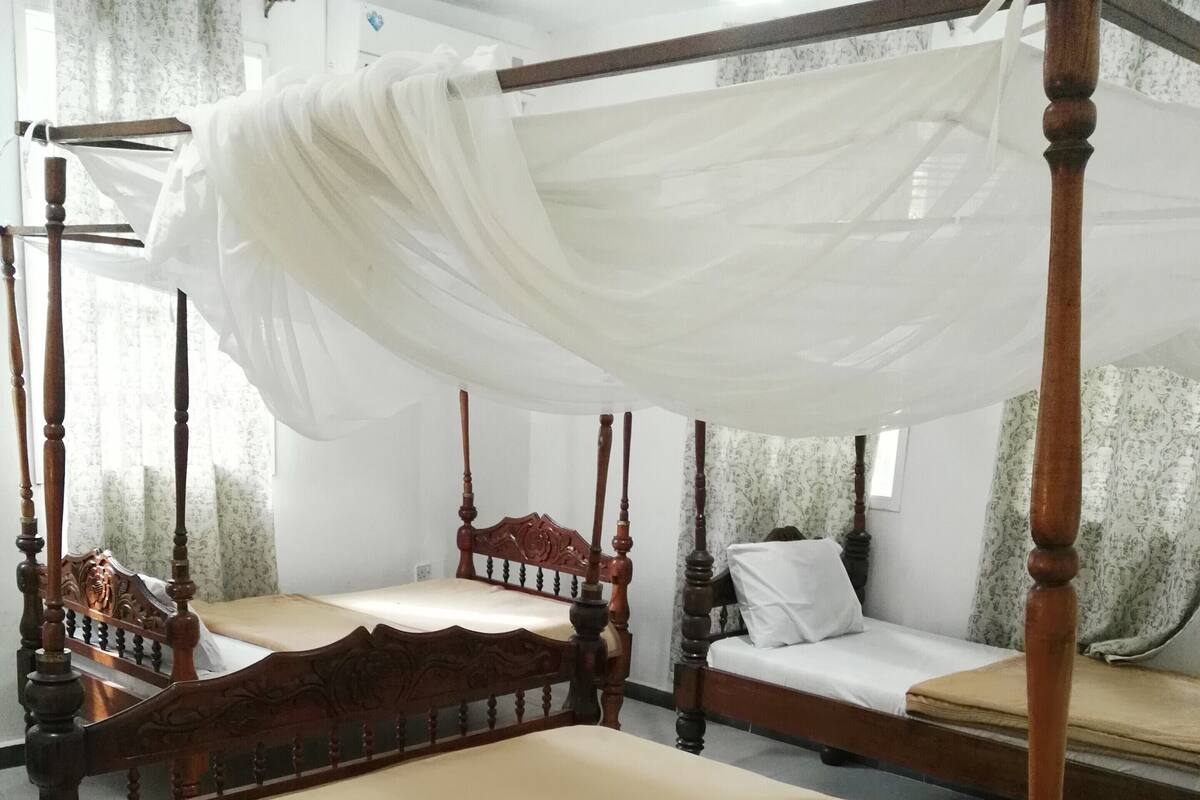 A traditional four-poster bed with a canopy, a smaller single bed, and a double bed in a room with white walls and curtains.