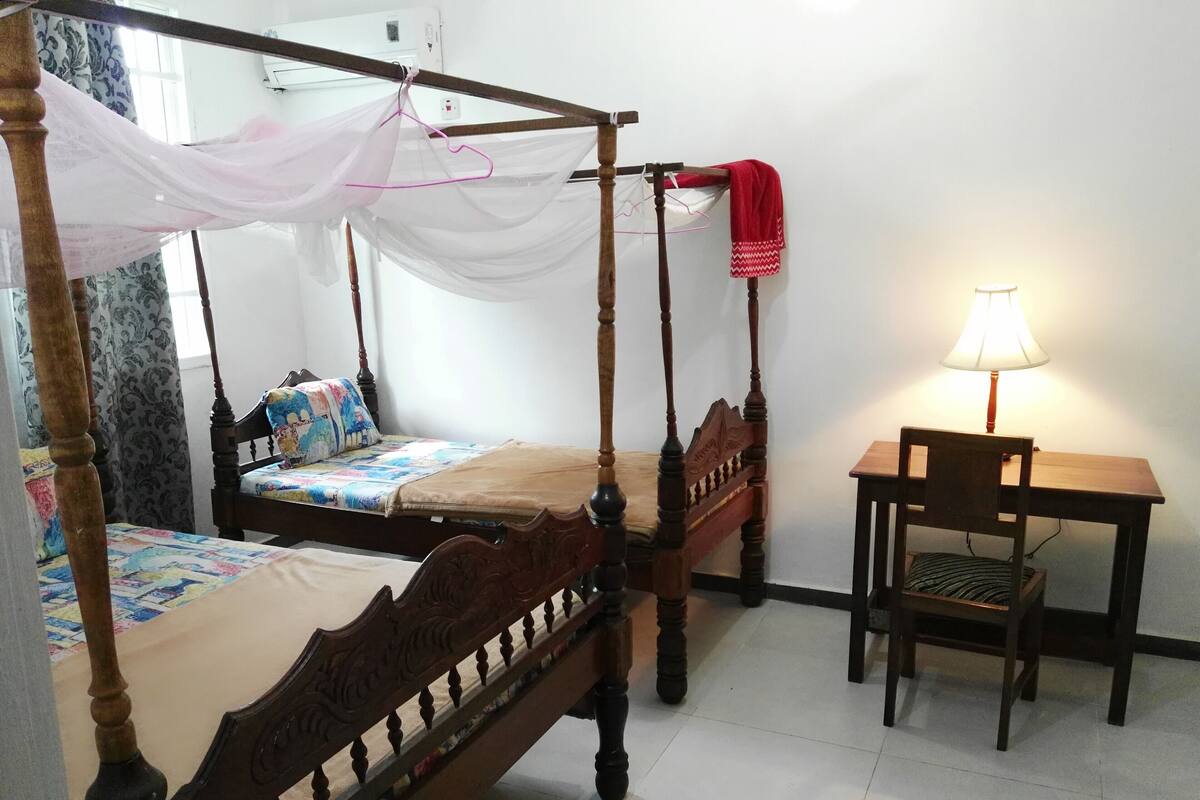 A bedroom with two wooden four-poster beds, a small wooden table with a chair, and a bedside lamp.
