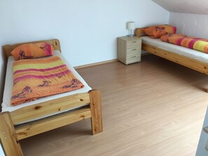 Une chambre avec deux lits superposés en bois, chacun étant équipé d’un matelas et d’une couverture. Il y a une table de chevet avec une lampe sur un des lits et une petite commode sur l’autre. La chambre a un plancher en bois et des murs blancs.