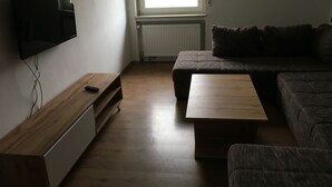 En stue med et fladskærms-tv på væggen, et træsofabord, en sofa og et vindue med naturligt lys.