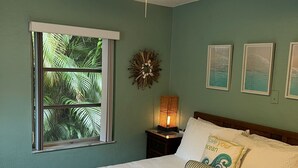 Une chambre avec un lit, un ventilateur de plafond, une fenêtre donnant sur des palmiers et des œuvres d’art murales.