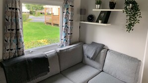 A modern living room with a grey sofa, a window with patterned curtains, a small shelf with decorative items, and a view of a garden. - Normandy: nature, history and comfort in a mobile home (Litteau)