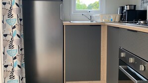 A compact kitchen with a stainless steel refrigerator, a built-in oven, and a window with a view of greenery. - Normandy: nature, history and comfort in a mobile home (Litteau)