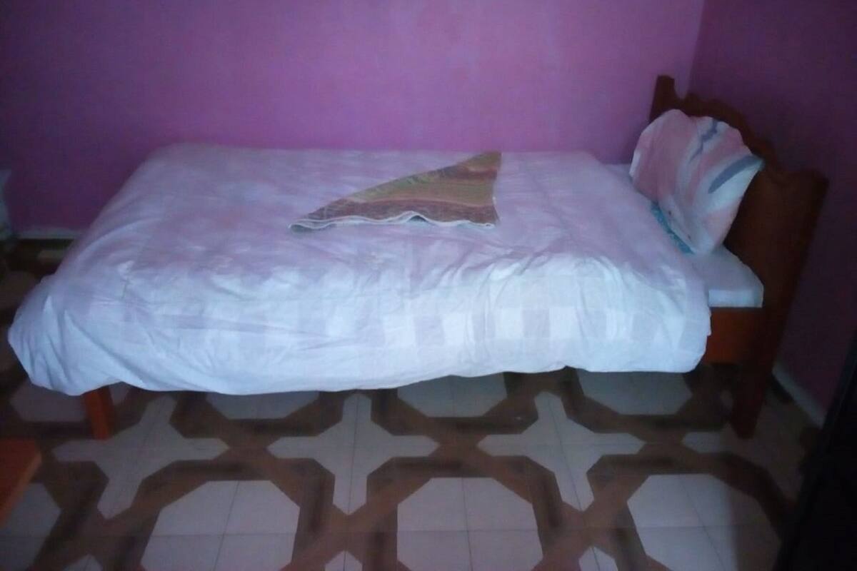 A single bed with a patterned bedspread and a decorative pillow in a room with a purple wall and a geometric floor design.