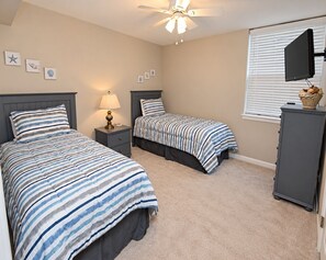 A bedroom with two beds, a nightstand with a lamp, a ceiling fan, a television, and a window with blinds.