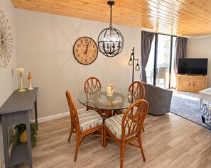 A dining area with a round glass table, four wicker chairs, a wall clock, a hanging light fixture, and a television in the background.