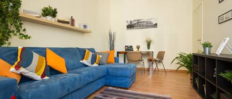 A living room with a blue sectional sofa, a striped rug, a desk with a chair, and a bookshelf with plants.