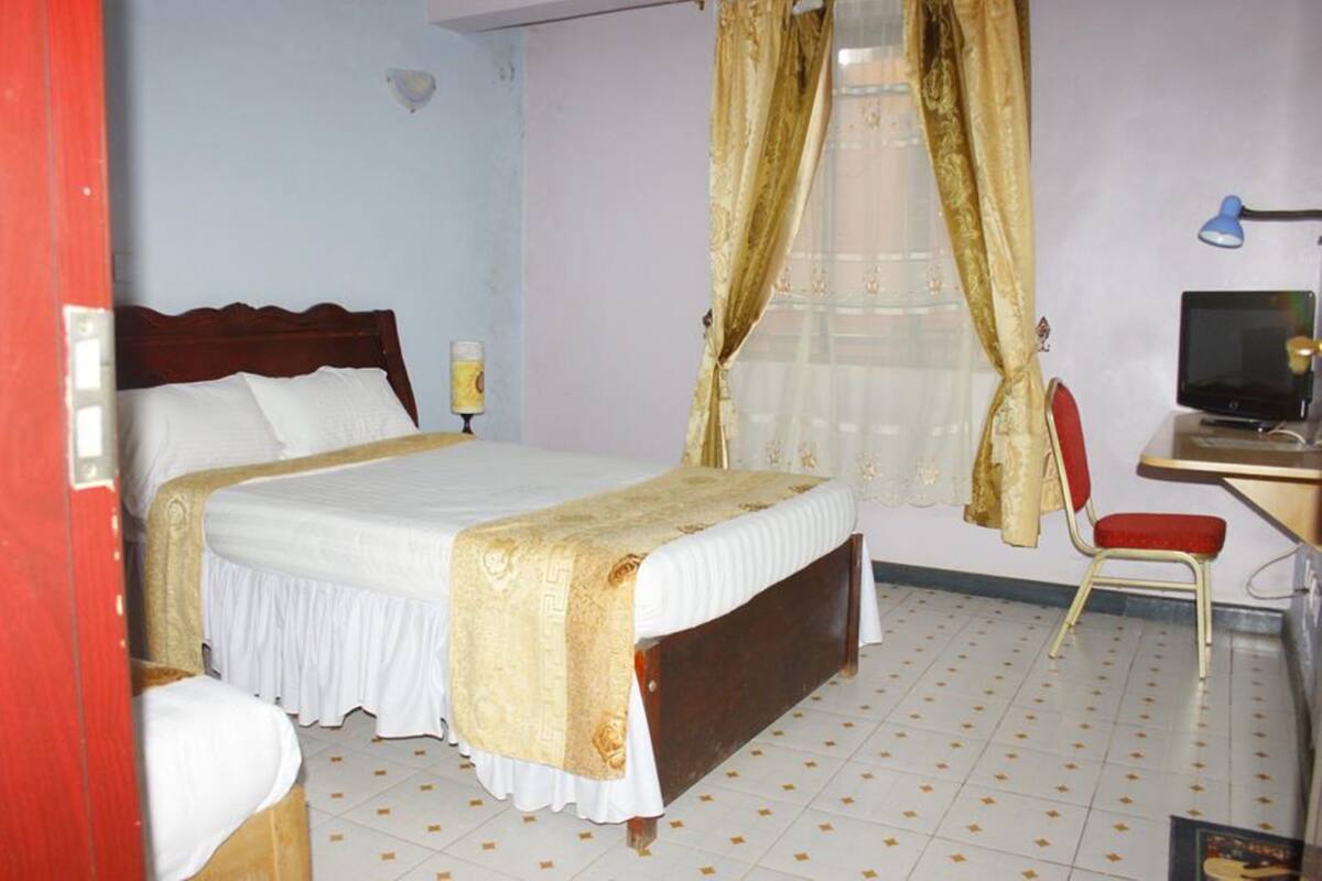 A bedroom with a bed, a desk with a computer, a chair, and a window with curtains.
