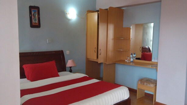 A bedroom with a bed, a wooden wardrobe, a small stool, a mirror, and a wall-mounted light.