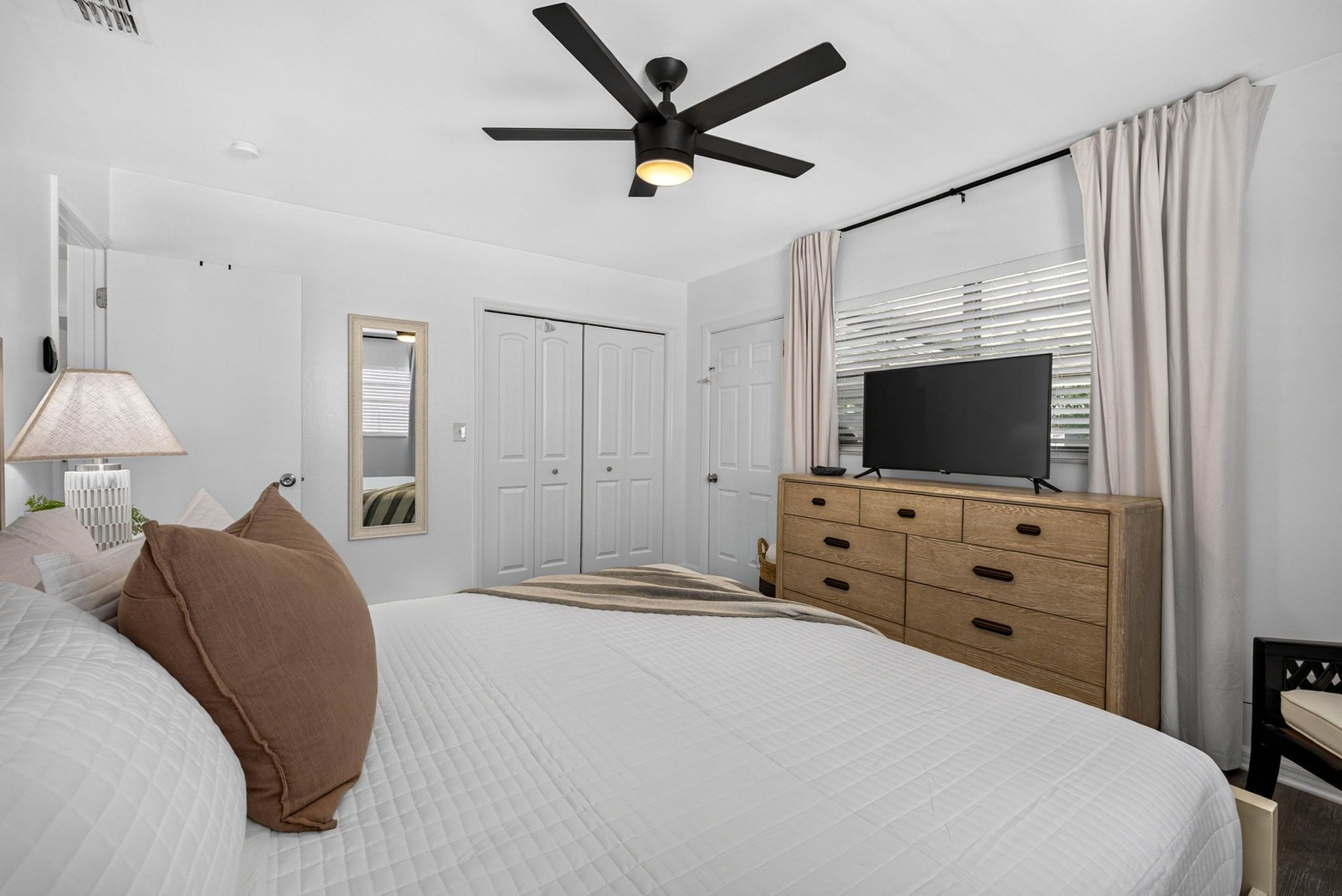 A bedroom with a bed, a dresser, a television, a ceiling fan, and a window with curtains.