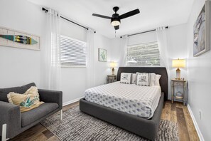Ein Schlafzimmer mit einem Bett, einem Schlafsofa, zwei Nachttischen mit Lampen, einem Deckenventilator und einem Fenster mit Jalousien.