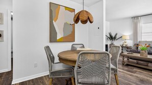 Een eethoek met een ronde houten tafel, vier rieten stoelen, een hanglamp en een modern abstract schilderij aan de muur.