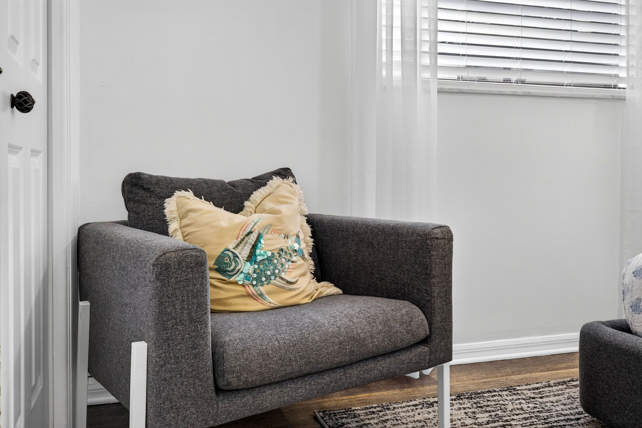 Un sillón gris con un cojín decorativo frente a una pared blanca, una ventana con persianas y un suelo de madera.