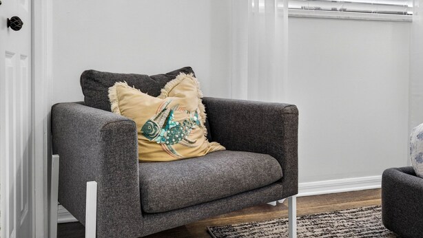 Een grijze fauteuil met een decoratief kussen tegen een witte muur, een raam met zonwering, en een houten vloer.
