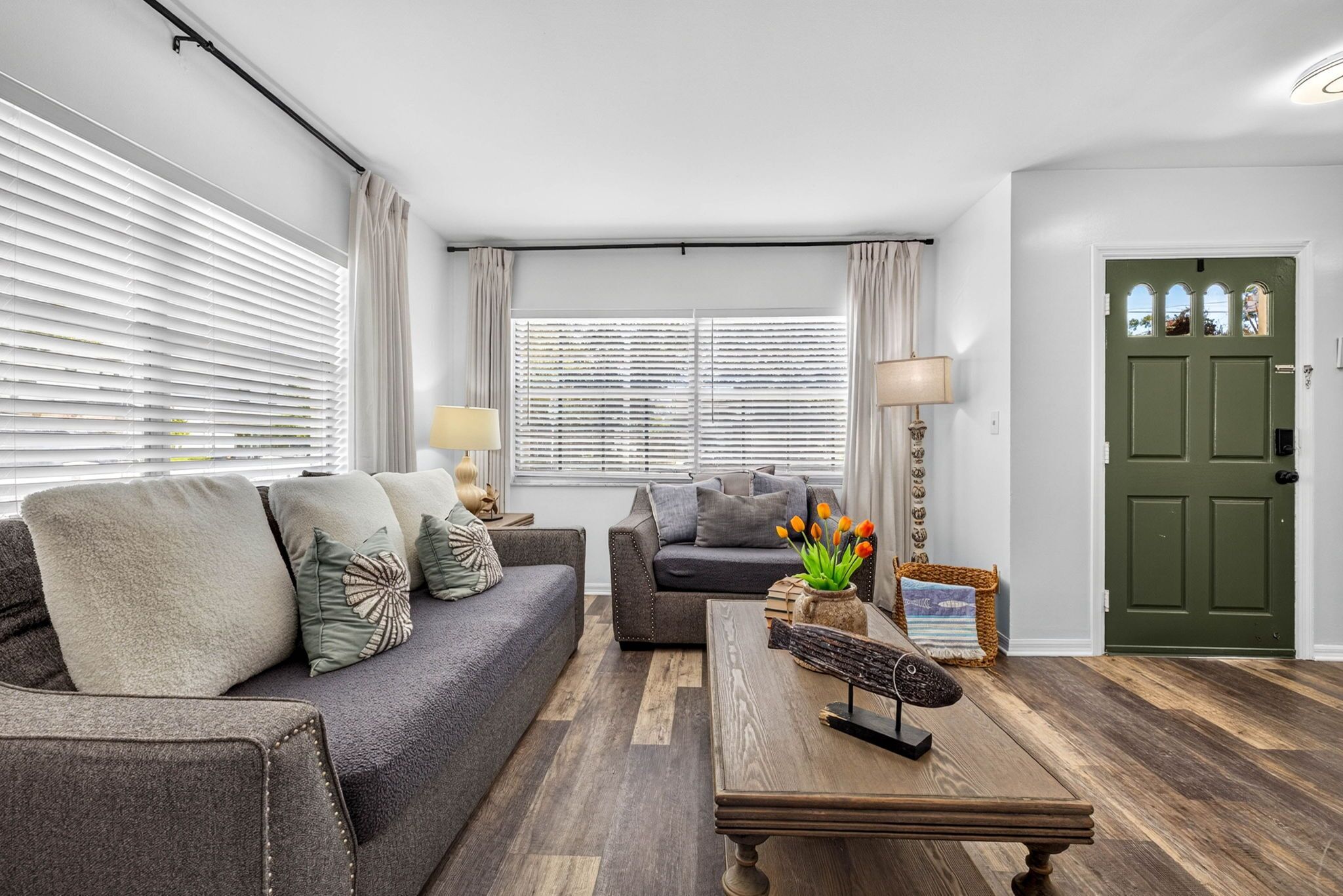Een woonkamer met een grijze bank, een kleinere grijze fauteuil, een houten salontafel en een lamp. Er is een deur met een groene omlijsting en een raam met zonwering.