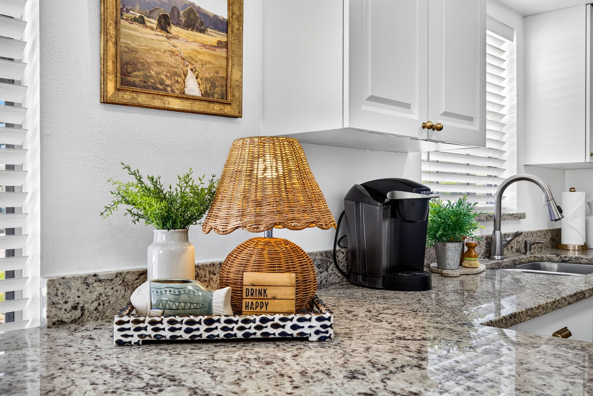 A kitchen counter with a coffee maker, a wicker lamp, a potted plant, a fish-shaped container, and a decorative sign.