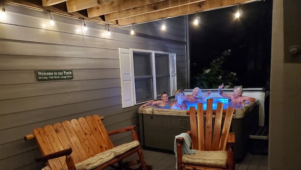 A wooden deck with two wooden chairs and a small table. A hot tub is visible in the background with people in it. - Maggie Valley Mountain View Suite & Spa (Maggie Valley)
