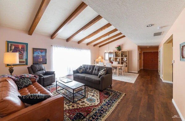 Amplia sala de estar con un sofá de cuero marrón, un sofá gris y una mesa de centro. El techo de madera exhibe vigas a la vista, una ventana grande con cortinas y una estantería con objetos decorativos.