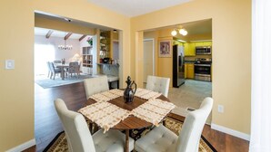A dining area with a wooden table, white chairs, and a chandelier. There is a kitchen visible in the background with yellow cabinets and stainless steel appliances.
