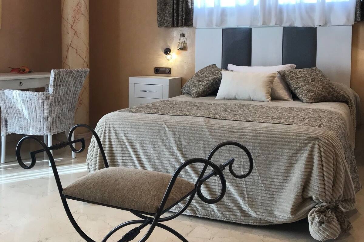 A bedroom with a bed, a nightstand, a chair, a window with curtains, and a wall-mounted light fixture.