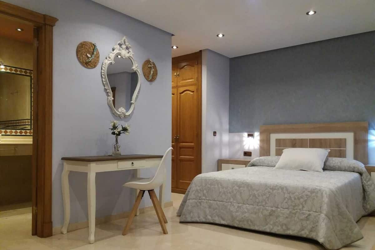 A bedroom with a bed, a desk, a chair, a mirror, and a door.