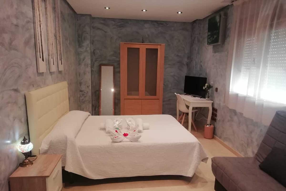 A bedroom with a bed, bedside table, a desk with a chair, a television, and a sofa.