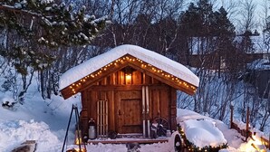 Une cabane en bois, dont le toit est recouvert de neige, et dont la véranda est illuminée.