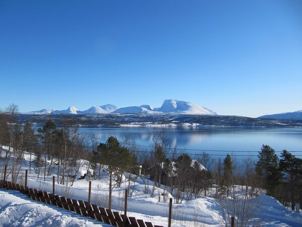 A snowy landscape with a lake, mountains, and a wooden fence in the foreground.
