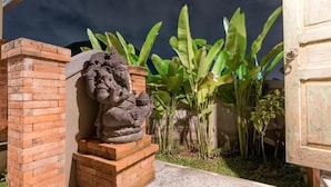 Een stenen beeld van een figuur op een bakstenen sokkel, omringd door weelderige groene bananenplanten.
