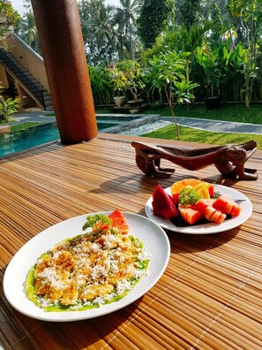 A dining table with a plate of food and a plate of fruit, set against a backdrop of a pool and tropical plants. - Umah Dangin Guesthouse - Hostel (Munggu)