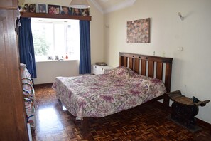 A bedroom with a wooden bed, a window with curtains, a small bedside table, and a wall-mounted painting. - Bustani Apartment (Mombasa)