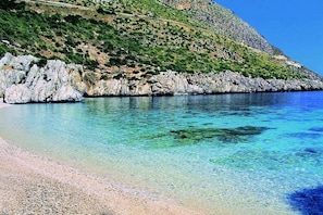 A coastal beach with clear turquoise water, rocky cliffs, and a pebbly shoreline.