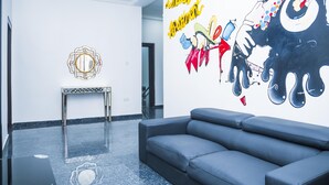 A lobby with a grey sofa, a small table, and a wall with a mirror and a colorful mural. - Lineech Apartments (Abuja)