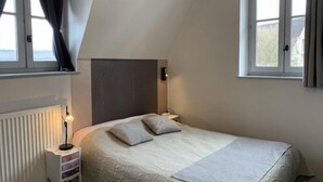 A bedroom with a bed, two pillows, a bedside lamp, a radiator, and two windows with curtains.