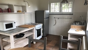 Cozinha compacta com fogão a gás, micro-ondas e frigorífico. Há uma mesa de madeira e cadeiras, e uma janela que proporciona luz natural.