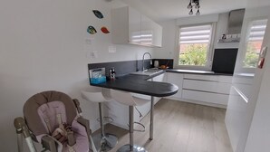 Eine moderne Küche mit weißen Schränken, einer schwarzen Arbeitsplatte und einem Fenster mit Jalousien. Zwei Barhocker und ein Rollstuhl stehen in der Ecke.
