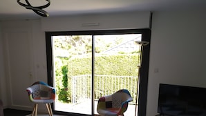 Ein Zimmer mit einer großen Glasschiebetür, die auf einen Balkon mit weißem Zaun und viel Grün führt. Zwei stilisierte Stühle und eine Deckenleuchte sind ebenfalls zu sehen.