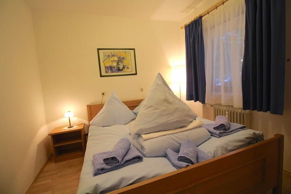 A single bed with pillows and towels, a bedside table with a lamp, a framed picture on the wall, and a window with curtains.