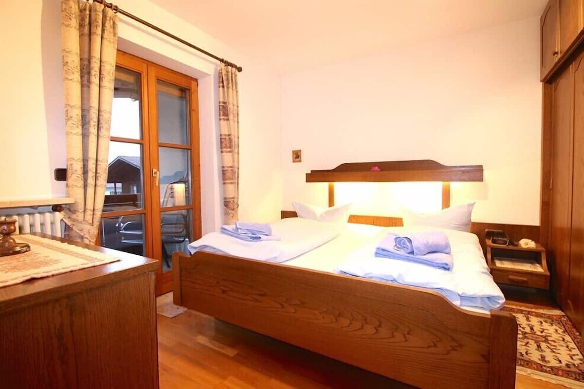 A bedroom with a wooden bed, a bedside table, a nightstand, a lamp, and a window with curtains.