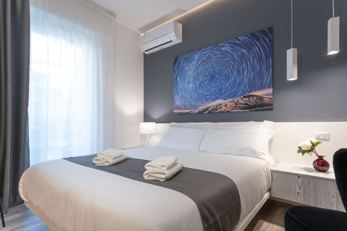 A modern hotel room with a large bed, a night sky mural, a bedside table with a vase, and an air conditioning unit.