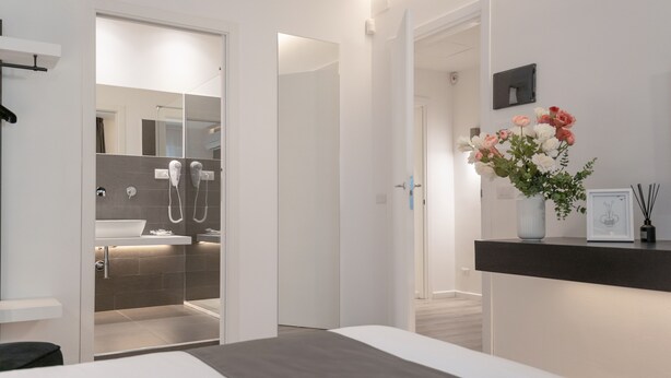 Ein modernes Hotelzimmer mit einem großen Bett, einem Badezimmer mit Dusche und Waschbecken sowie einer kleinen Schreibtischzeile mit einem Blumenstrauß.